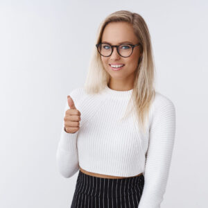 Nice work, proud of you. Pleased and delighted attractive elegant and flirty woman in glasses and skinny white sweater showing thumbs up gesture and smiling impressed against white background.